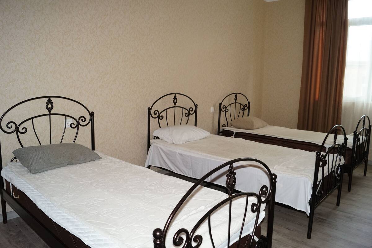 Three single beds with metal frames in a room with plain walls and a window with curtains.
