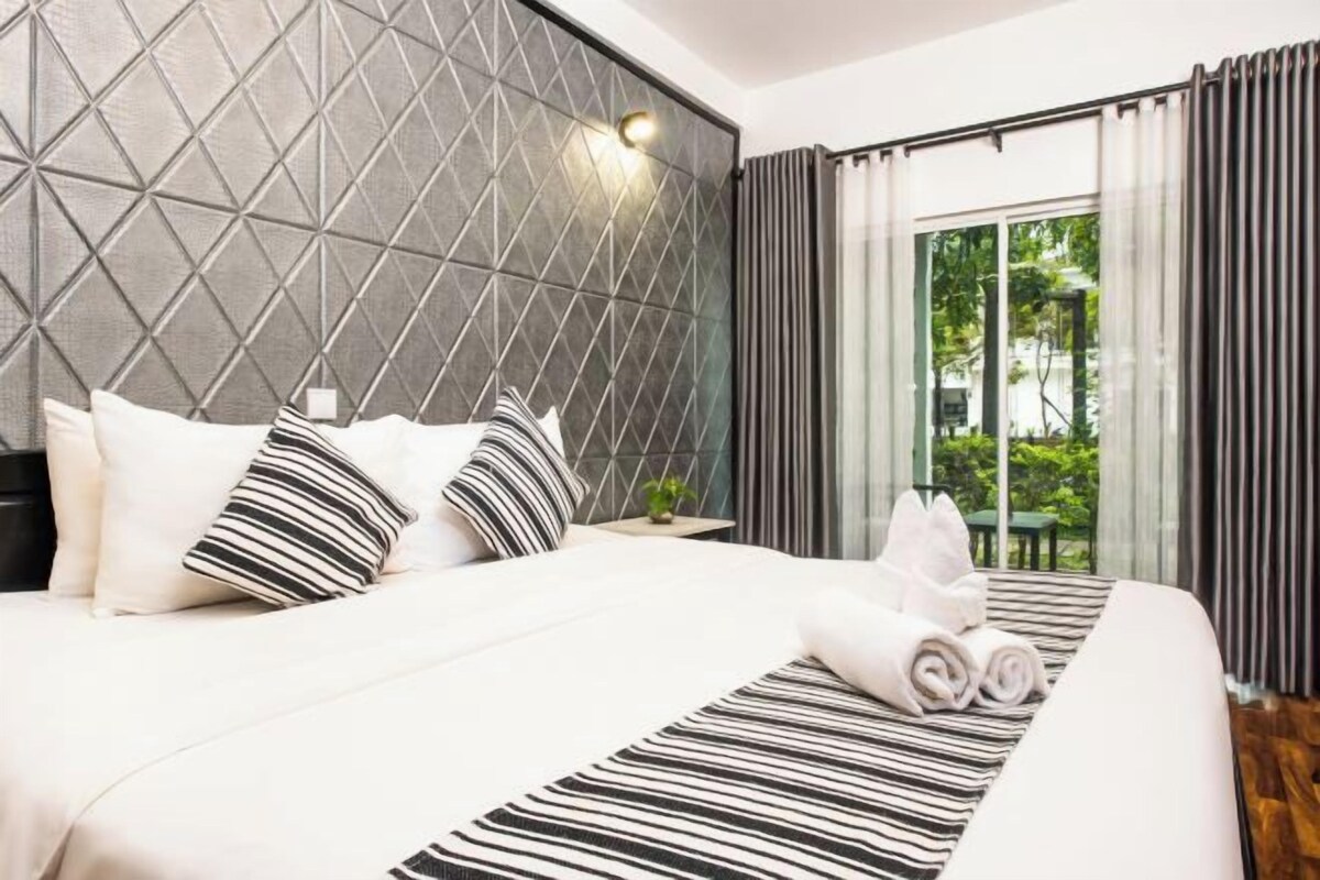 A modern hotel room with a geometric-patterned headboard, a neatly made bed with striped and plain pillows, and a view of greenery through large windows.