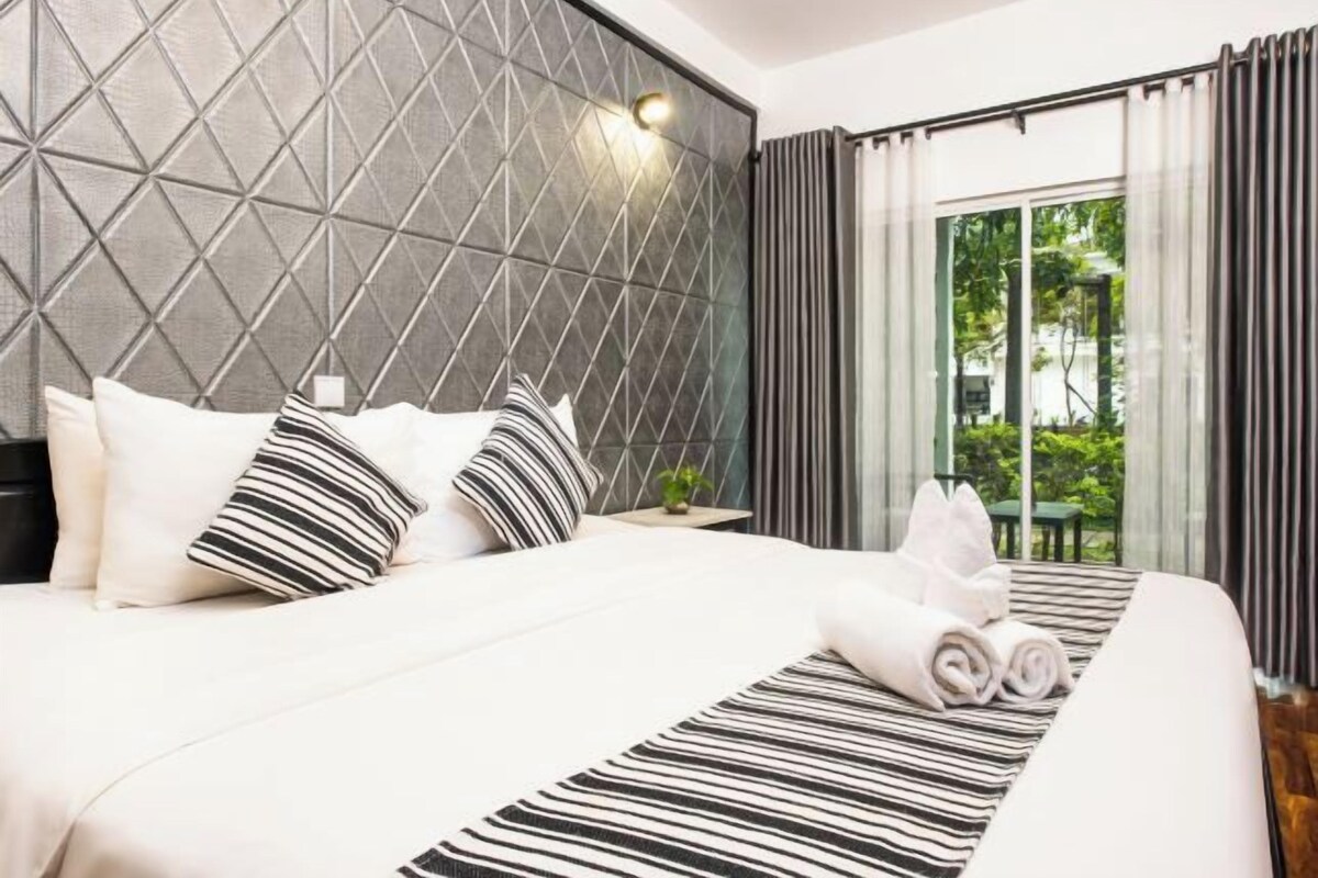 Ein modernes Hotelzimmer mit einem geometrisch gemusterten Kopfteil, einem ordentlich bezogenen Bett mit gestreiften und einfarbigen Kissen und Blick auf Grünanlagen durch große Fenster.