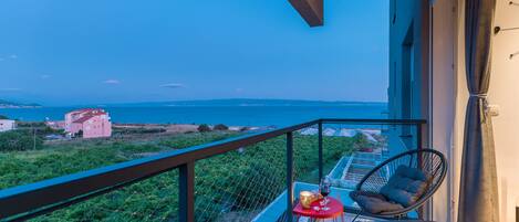 A balcony with a view of the sea, a chair, a small table with a glass of wine, and a red tablecloth.