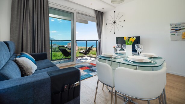 A modern living room with a glass dining table, white chairs, a grey sofa, and a large window with a view of the sea. - StellaMare Apartments (Split)