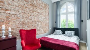 A bedroom with a brick wall, a large window with curtains, a red chair, and a bed with patterned pillows. - Elite Apartments Loft Center (Gdansk)