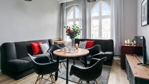 A modern living room with a black sofa, red and grey cushions, a round wooden table, and black chairs. There are two windows with curtains, a black pendant light, and a TV on a wooden stand. - Elite Apartments Loft Center (Gdansk)