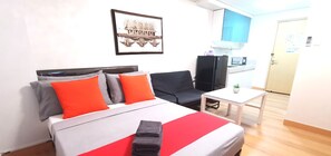 A hotel room with a bed, a desk, a chair, a small table, a picture on the wall, and a kitchenette area. - Butler's BnB (Quezon City)