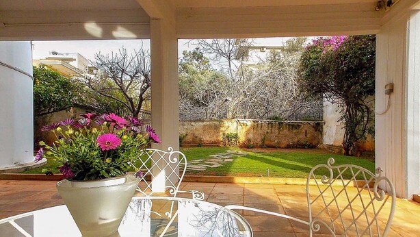 A glass table with a white chair, a potted plant with pink flowers, and a view of a garden with a lawn and trees. - Glyfada Spacious Apartment Garden (Glyfada)