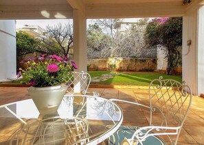 A glass table with a white chair, a potted plant with pink flowers, and a view of a garden with a lawn and trees. - Glyfada Spacious Apartment Garden (Glyfada)