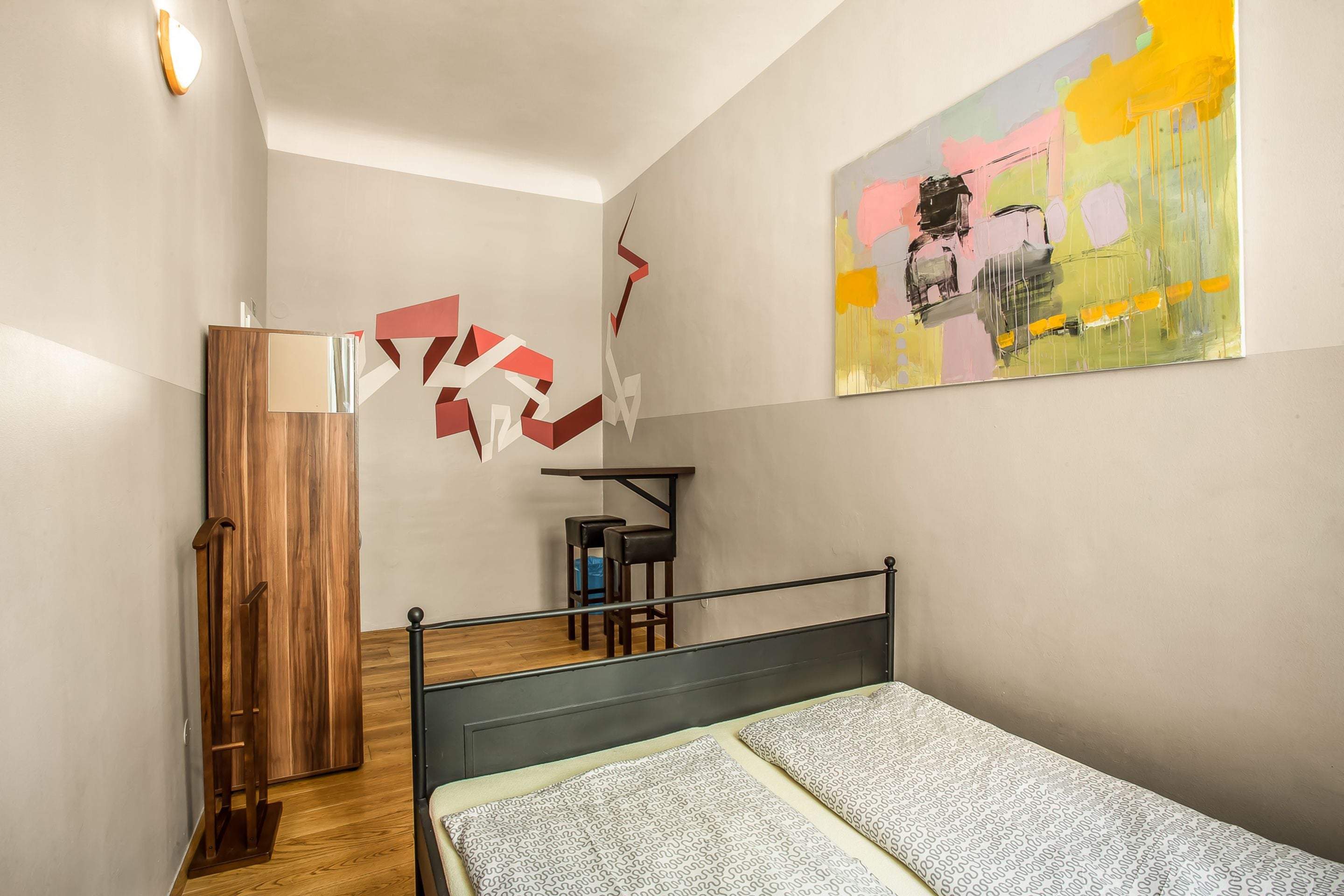 A bedroom with a bed, a wooden wardrobe, a desk with a chair, and a colorful abstract painting on the wall.