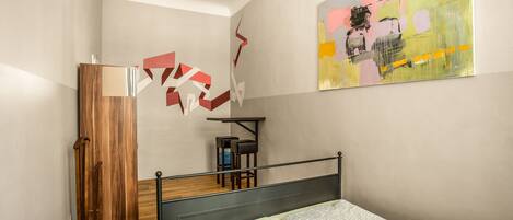 A bedroom with a bed, a wooden wardrobe, a desk with a chair, and a colorful abstract painting on the wall.