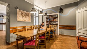 A dining area with a wooden table and chairs, a person working on a laptop, and a bookshelf with books and decorative items. - Ahoy! Hostel (Prague)