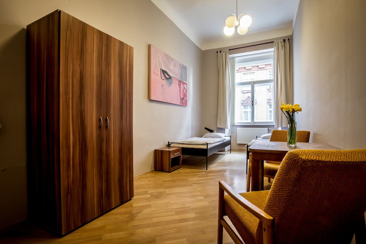 A room with a wooden wardrobe, a bed, a small table with a vase of flowers, and a window with curtains.