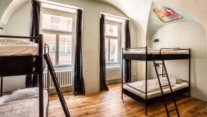 A dormitory room with bunk beds, wooden flooring, large windows with curtains, and a radiator. - Ahoy! Hostel (Prague)
