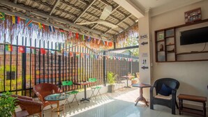 A sunlit patio with wicker chairs, a glass table, and a TV mounted on the wall. Flags are strung across the bamboo fence. - 333 HOSTEL, Siem Reap (Siem Reap)