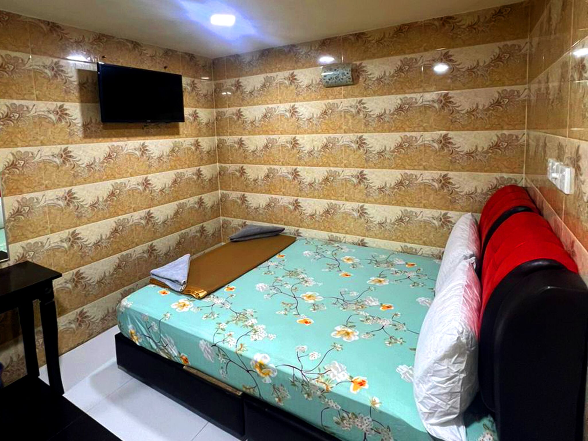 Una habitación pequeña y estrecha con papel tapiz floral, una cama con colcha floral, un televisor montado en la pared y una mesita.