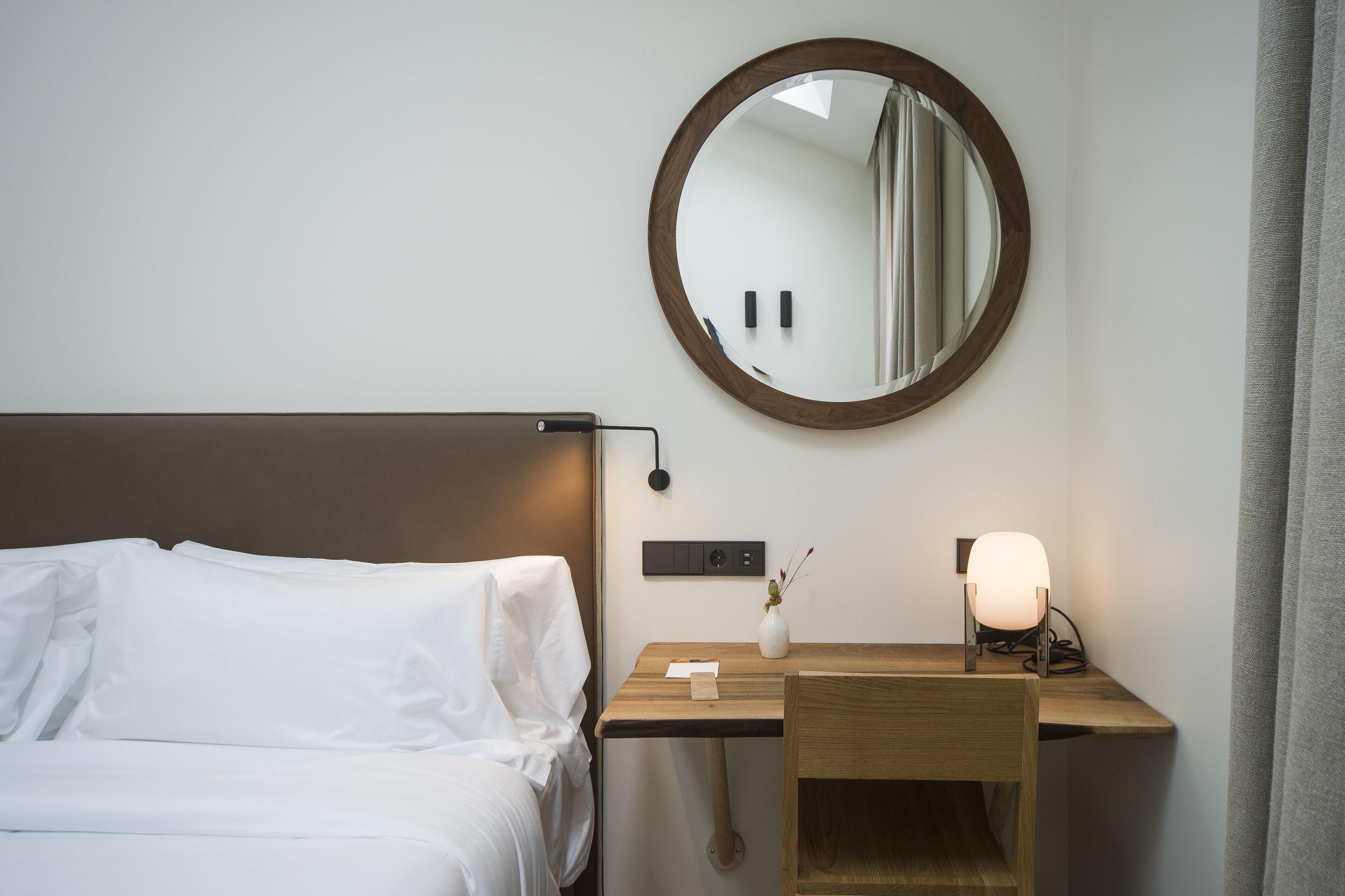 Een netjes opgemaakt bed met witte lakens, een nachtkastje met een lamp, een spiegel en een kleine vaas met een plantje.