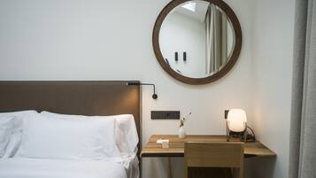 Een netjes opgemaakt bed met witte lakens, een nachtkastje met een lamp, een spiegel en een kleine vaas met een plantje.