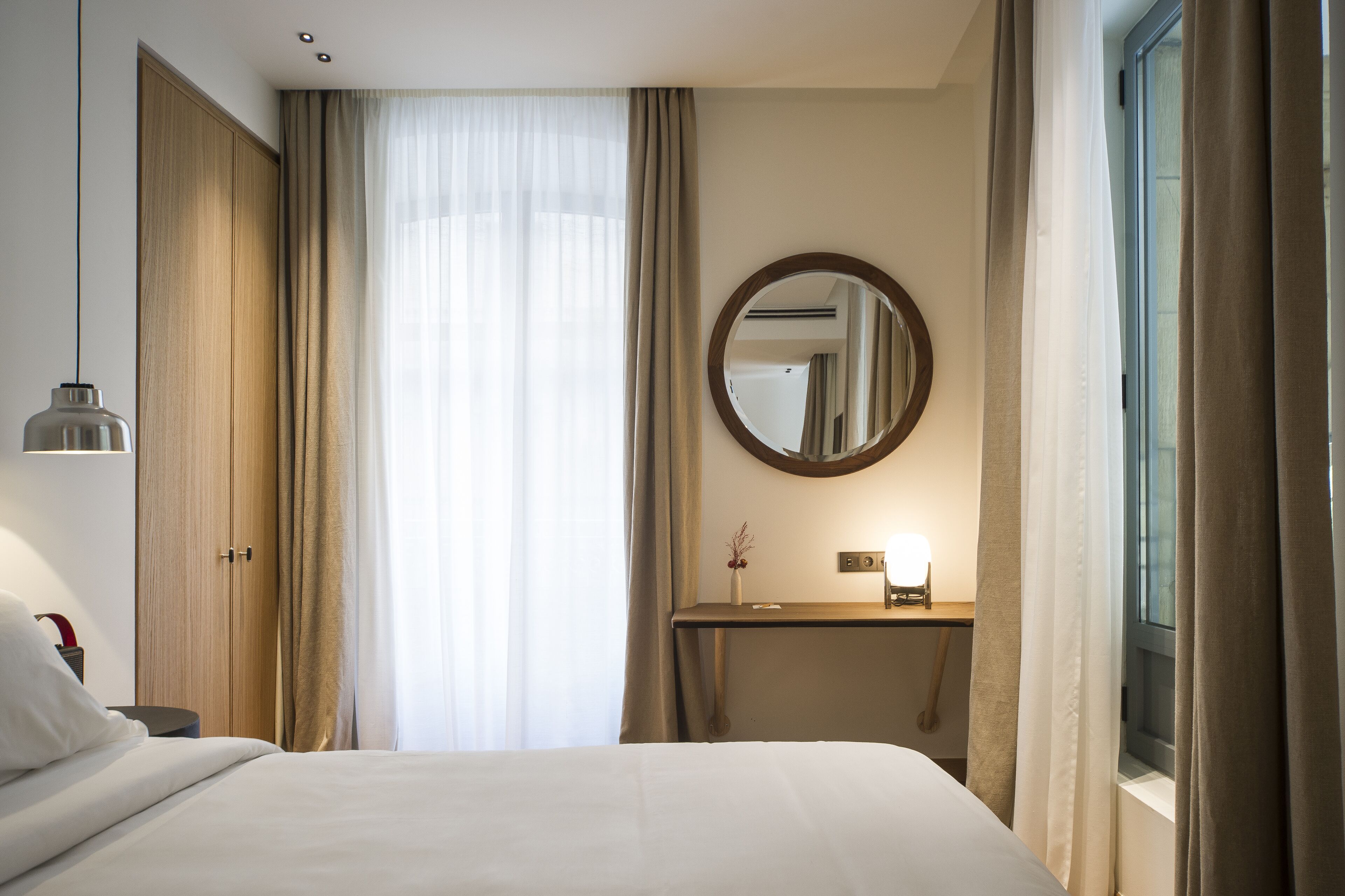 A hotel room with a bed, a bedside table, a mirror, and a window with curtains.