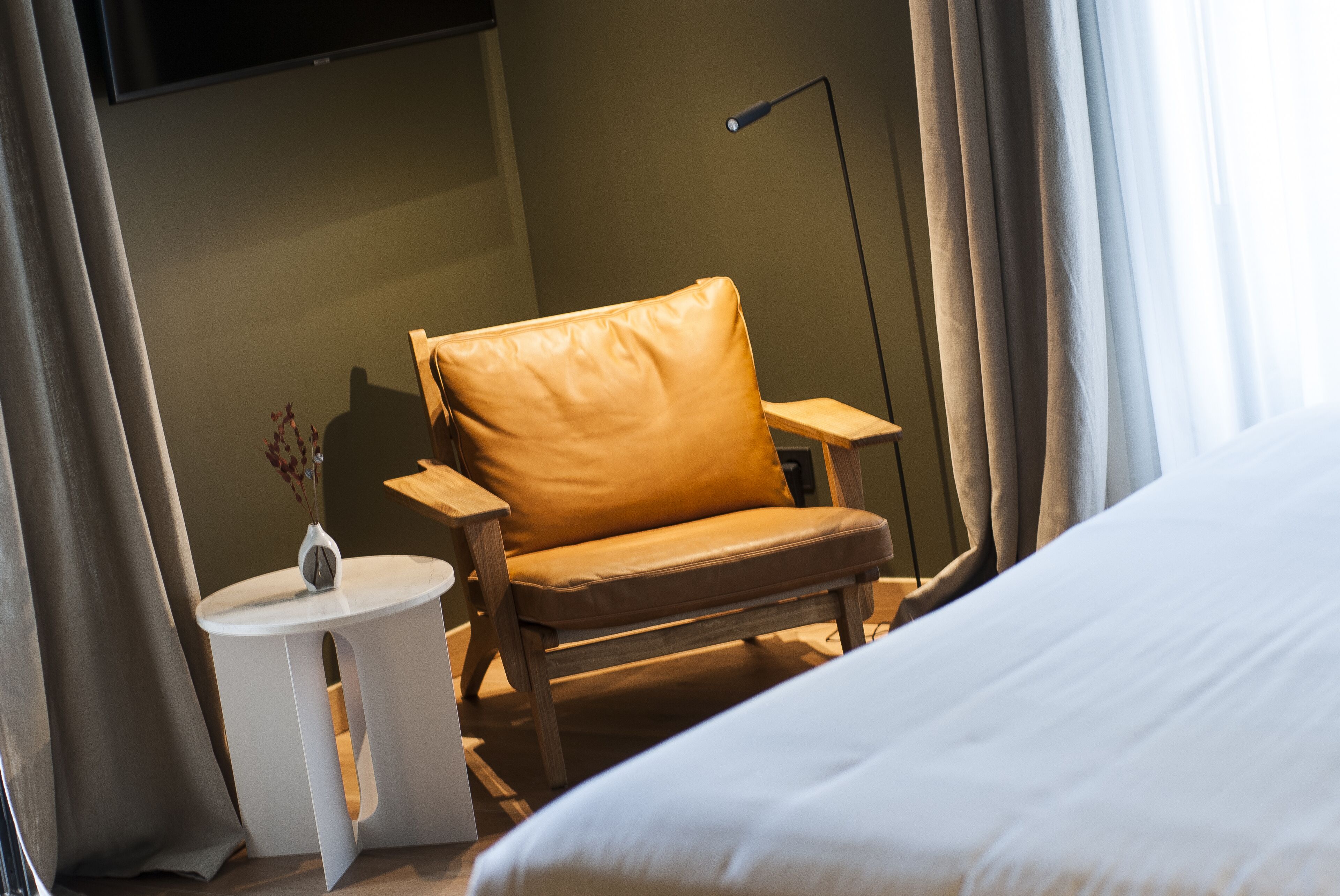 A hotel room with a bed, a wooden chair, a small round table, and a floor lamp.