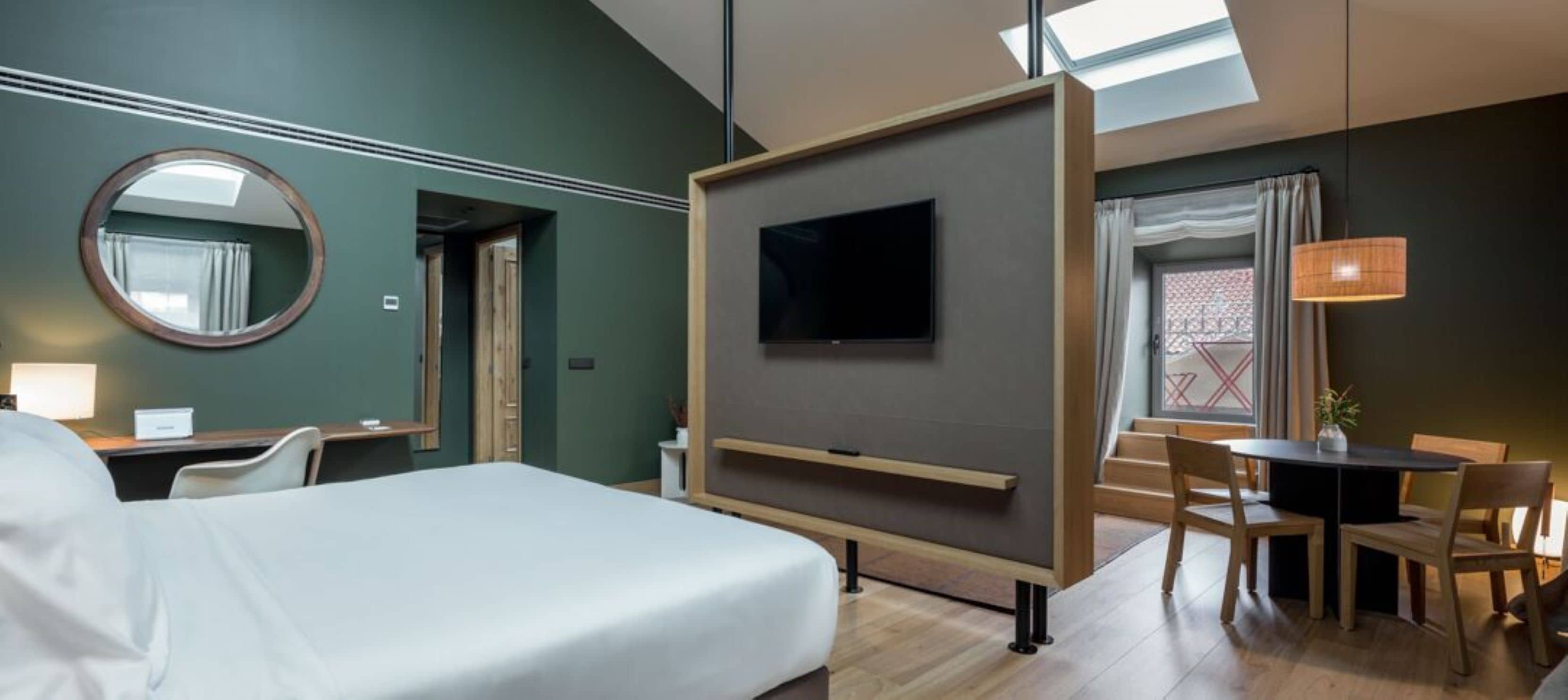 Una habitación de hotel con cama, televisor montado en la pared, escritorio con silla y zona de comedor con mesa y sillas.
