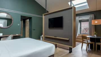 Una habitación de hotel con cama, televisor montado en la pared, escritorio con silla y zona de comedor con mesa y sillas.