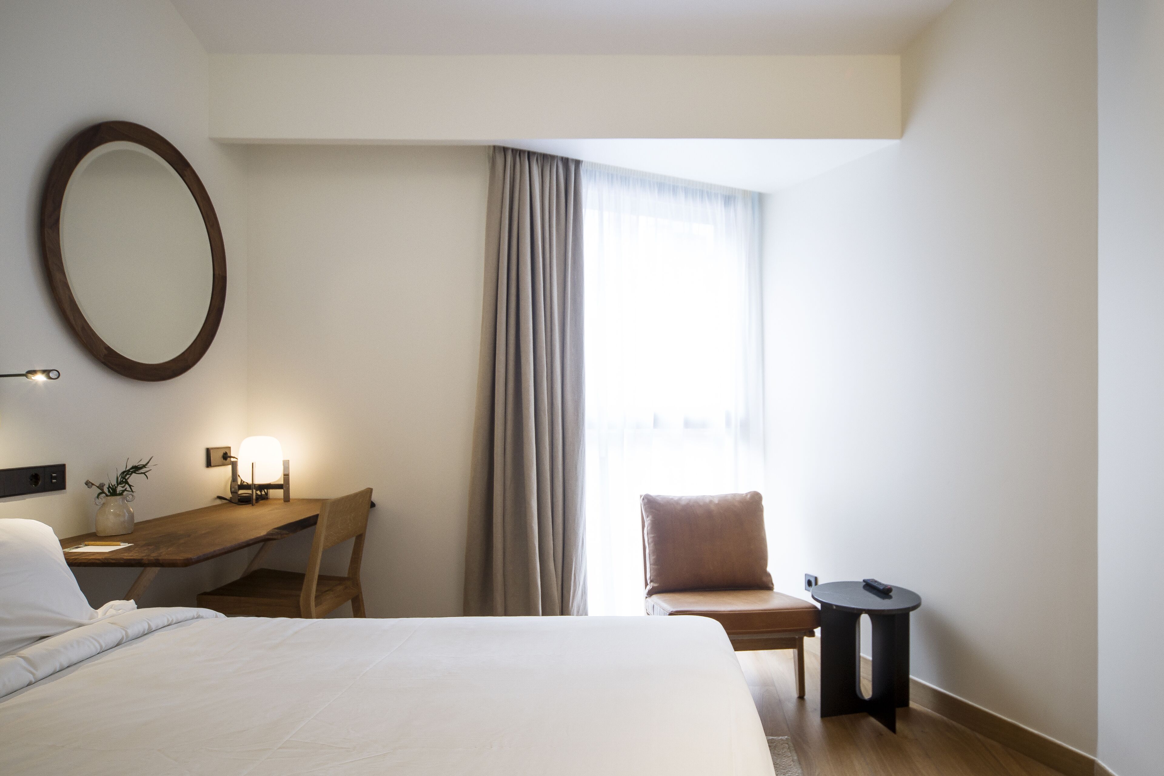 Habitación de hotel con cama, escritorio con silla, una mesita, un espejo redondo y una ventana con cortinas.