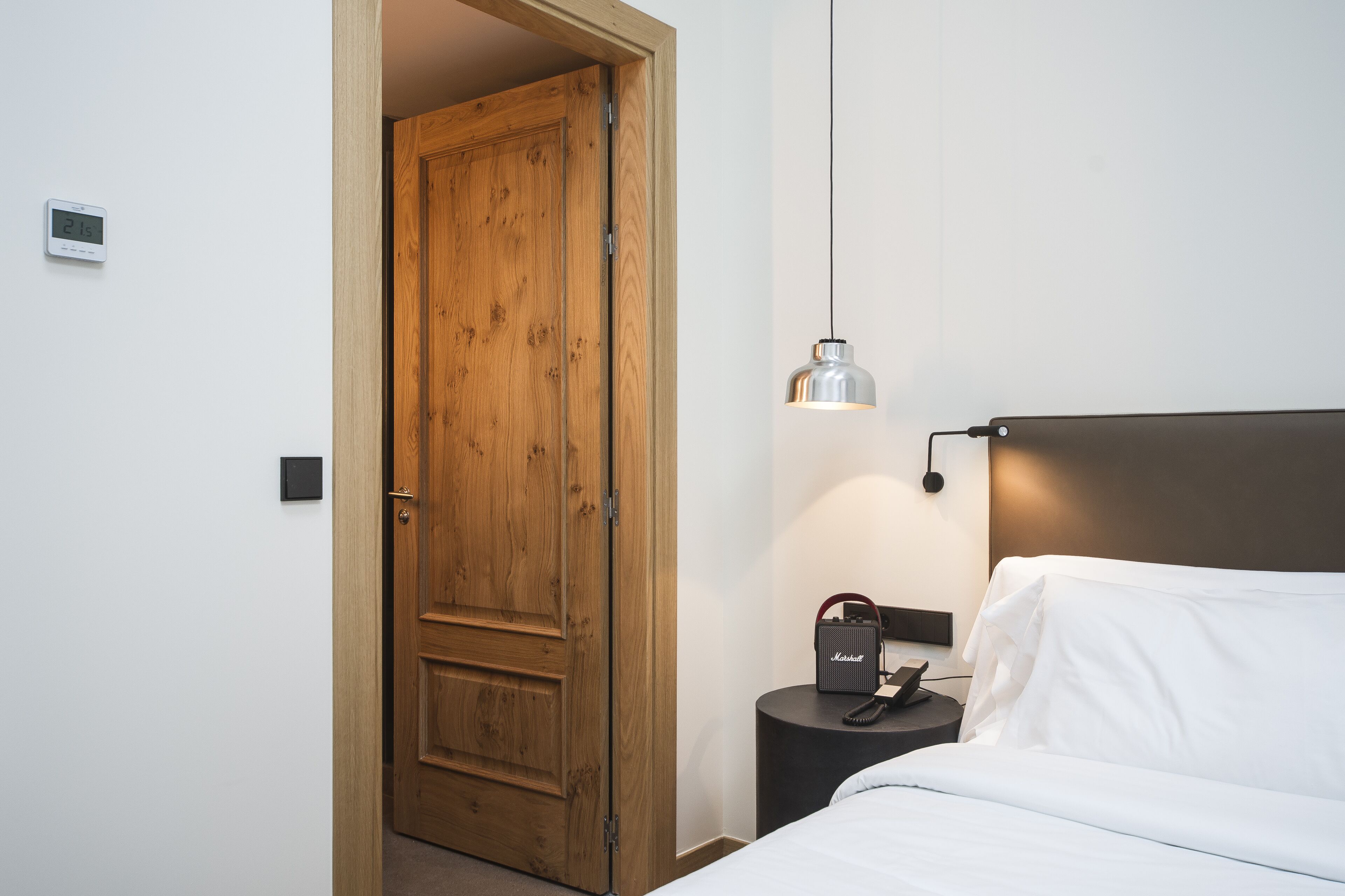 A hotel room with a wooden door, a bed with white bedding, a bedside table with a lamp, and a Marshall speaker.