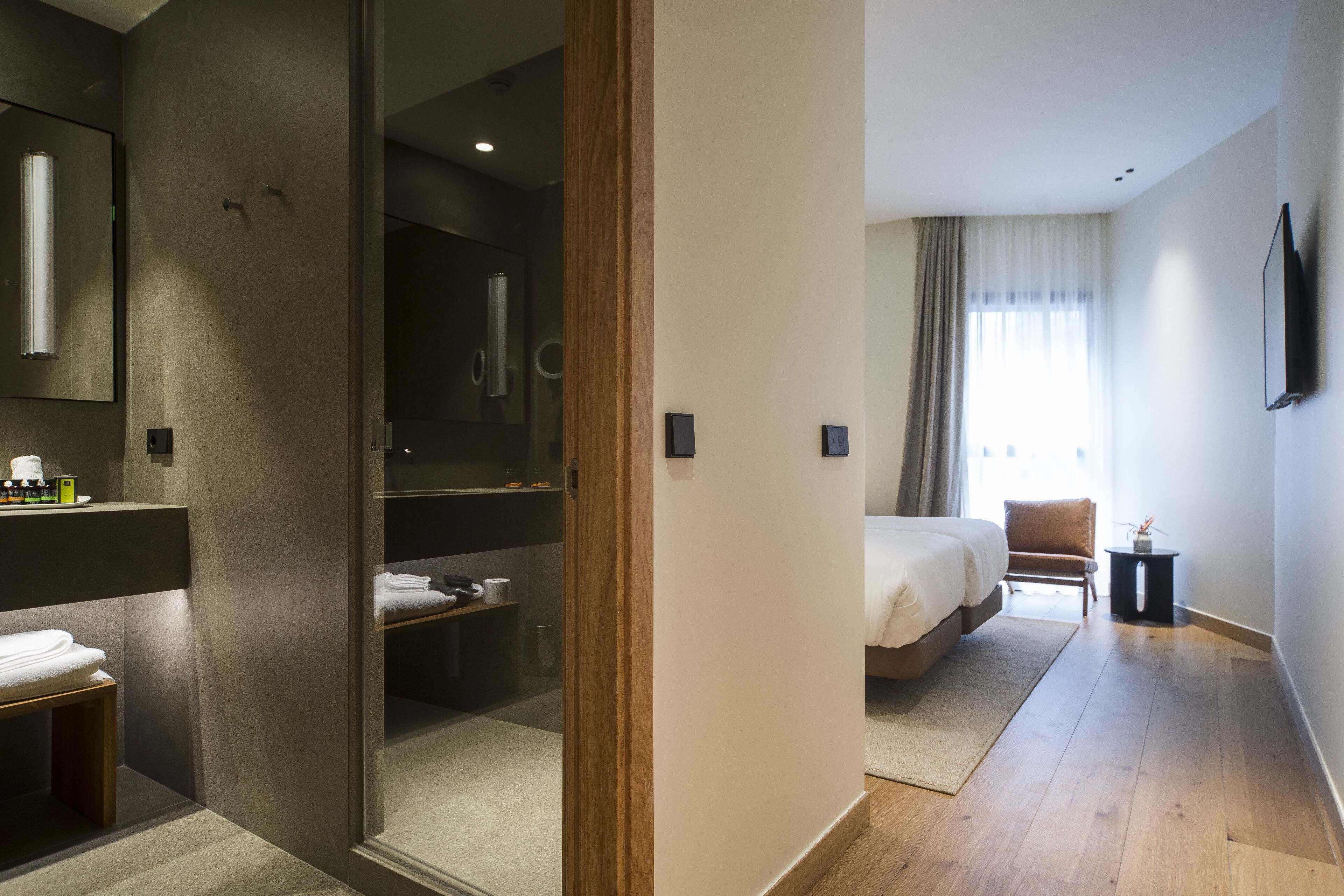 Una habitación de hotel moderna con una cama grande, una zona de estar con una silla, una mesita y un baño con cabina de ducha de vidrio.