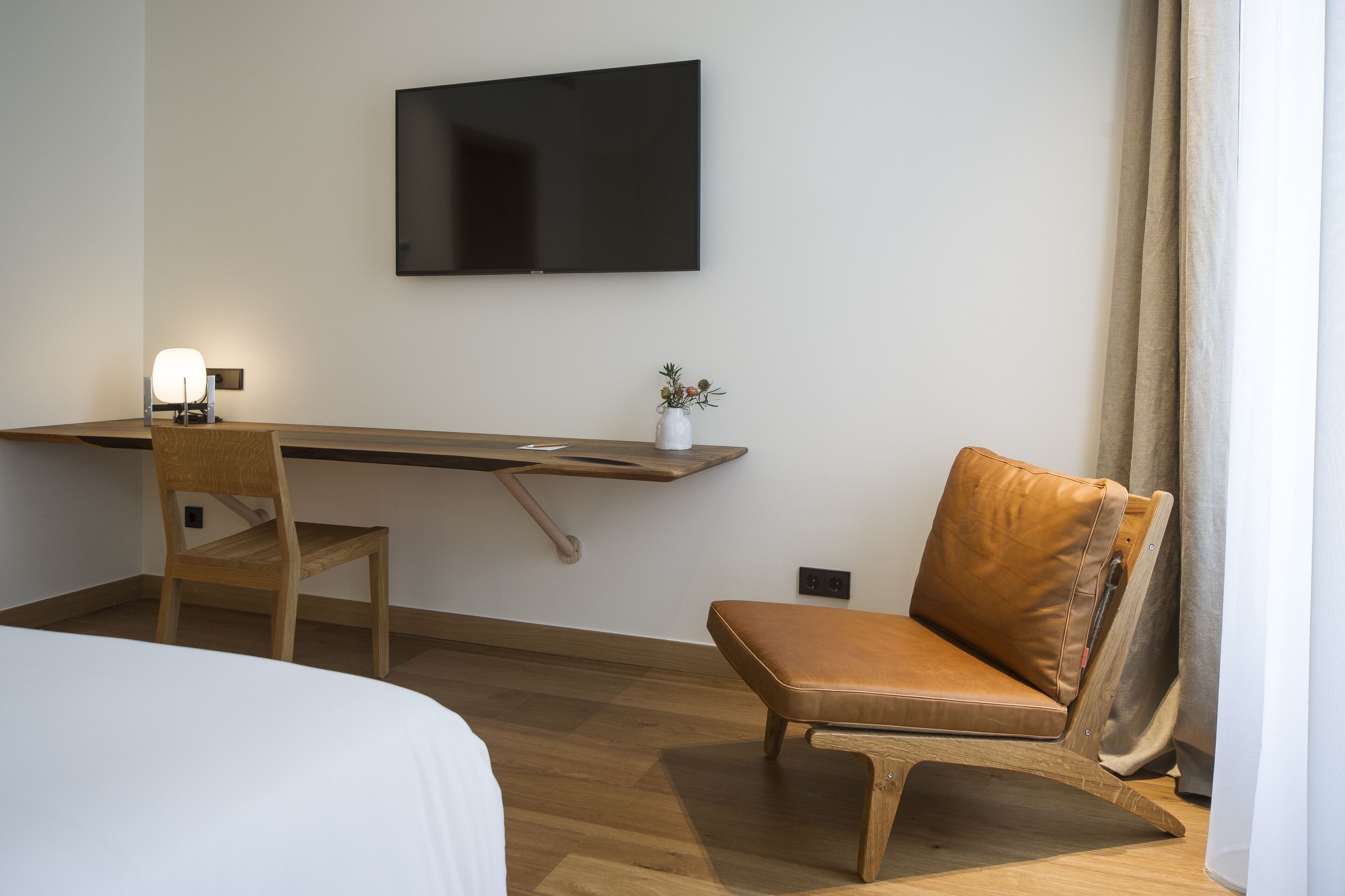 A hotel room with a bed, a desk with a chair, a television mounted on the wall, and a window with curtains.