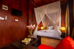 A room with a bed, a bedside table with a book and a vase of flowers, a television on the wall, and a red pillow on the chair. - Watdamnak House (Siem Reap)