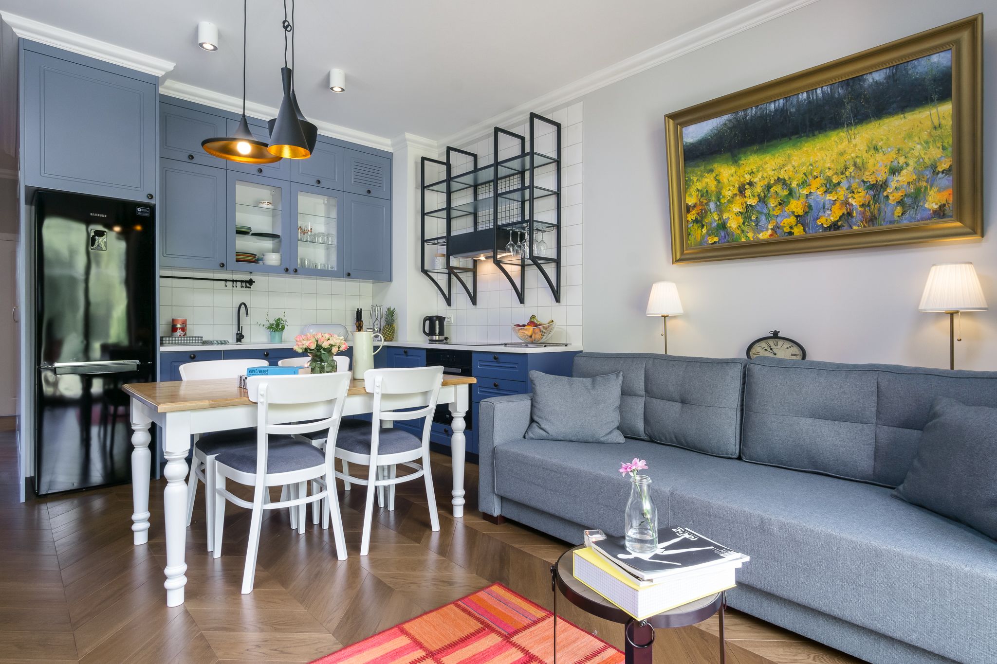 Una cocina moderna con armarios azules, una mesa de comedor con sillas, un sofá gris y un cuadro de un campo.