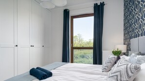 Ein Schlafzimmer mit Bett, Fenster mit Vorhängen, Nachttisch mit Lampe und einer Wand mit Muster.