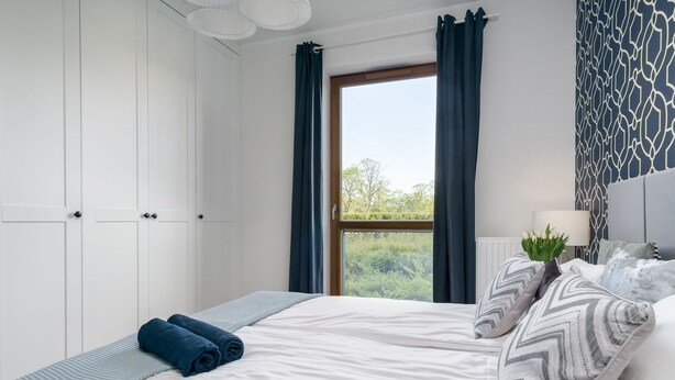 A bedroom with a bed, a window with curtains, a bedside table with a lamp, and a patterned wall.
