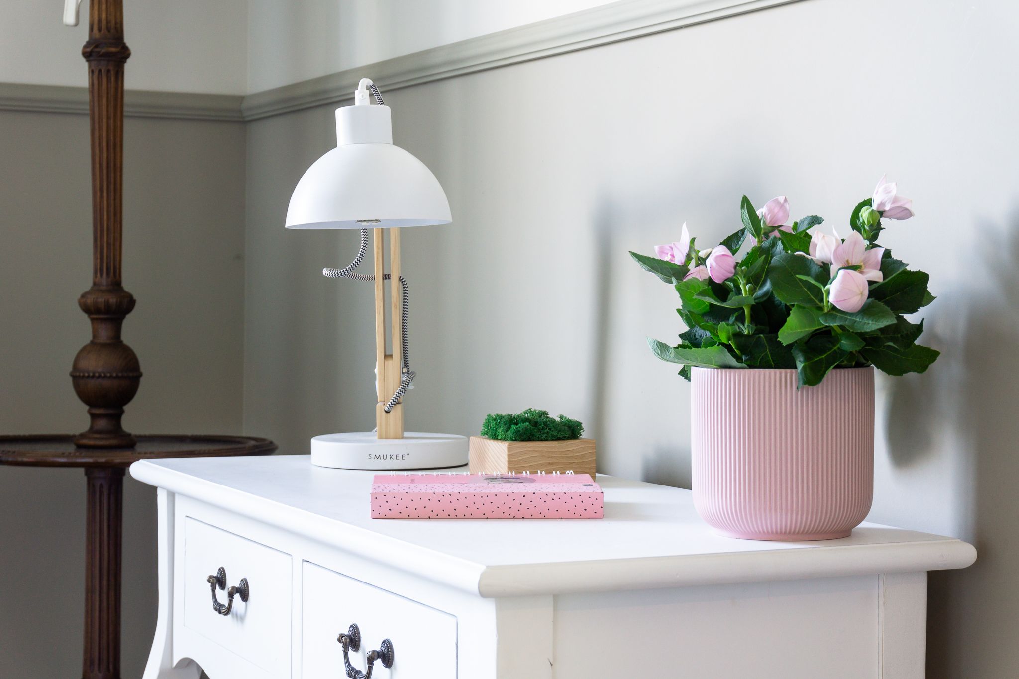 A white dresser with a white desk lamp, a pink polka-dotted notebook, a small wooden box, and a pink pot with green foliage.