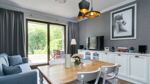 A modern living room with a wooden dining table, white chairs, a grey sofa, a TV, and a framed artwork on the wall.