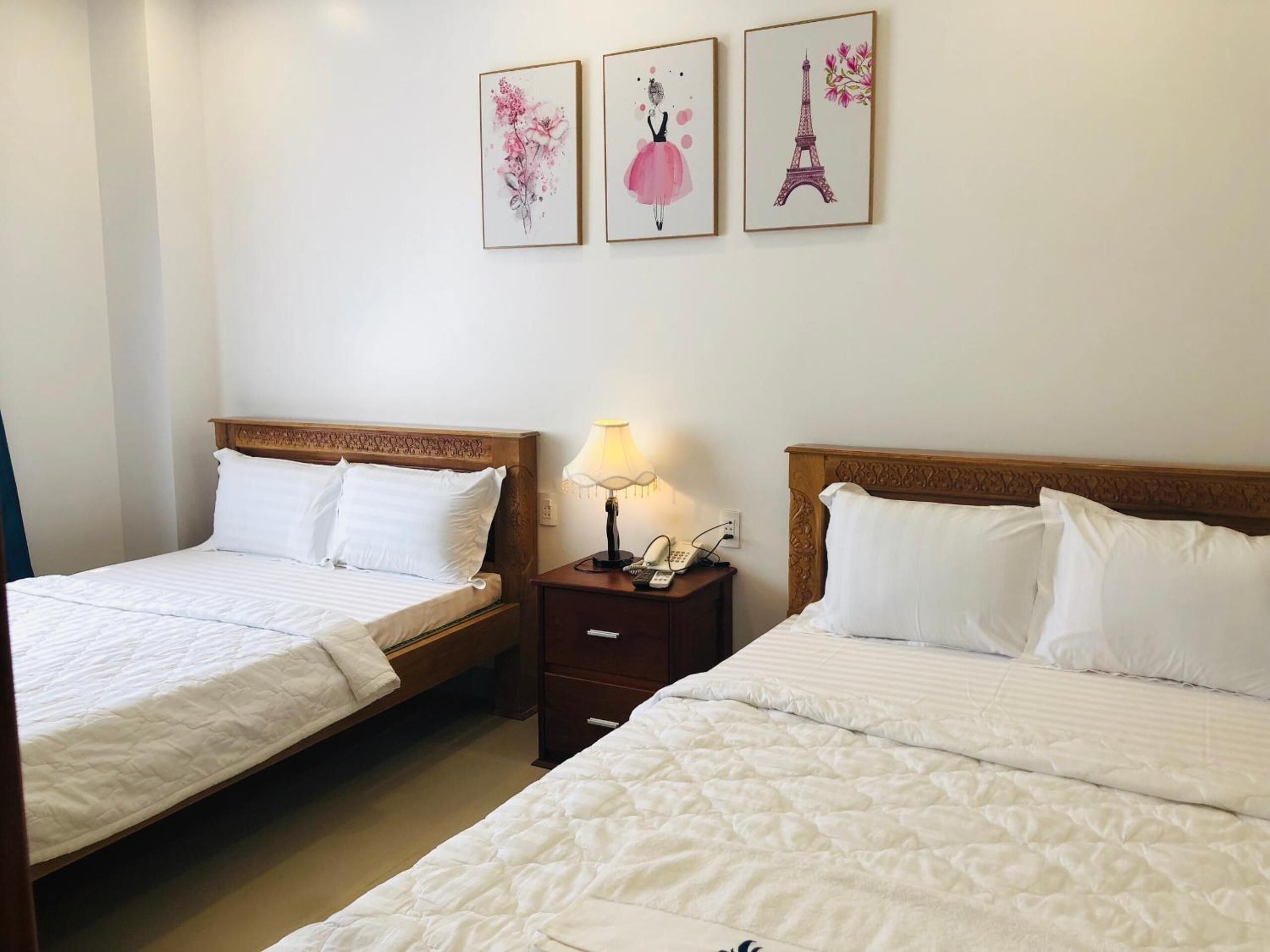 A hotel room with two beds, a nightstand with a lamp, and three framed artworks on the wall.