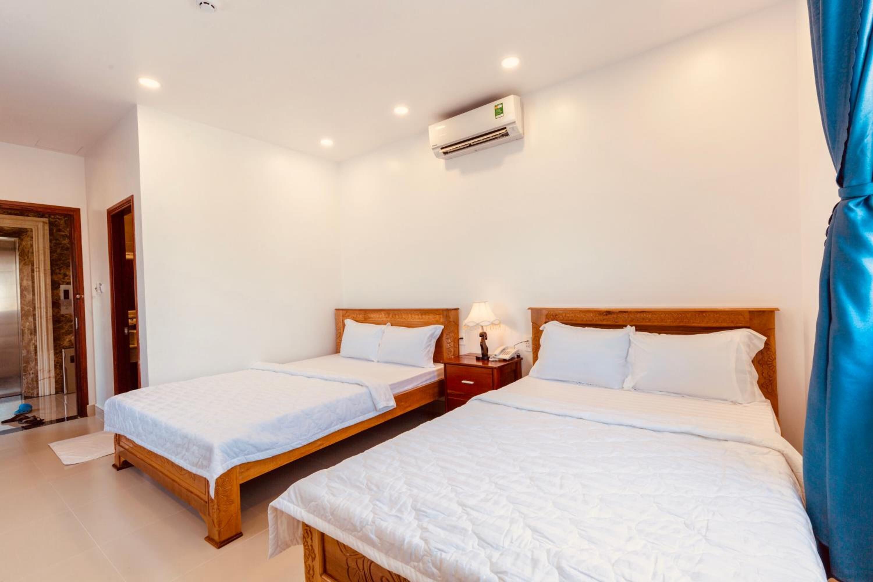 A hotel room with two single beds, a wooden headboard, a nightstand with a lamp, and an air conditioning unit on the wall.