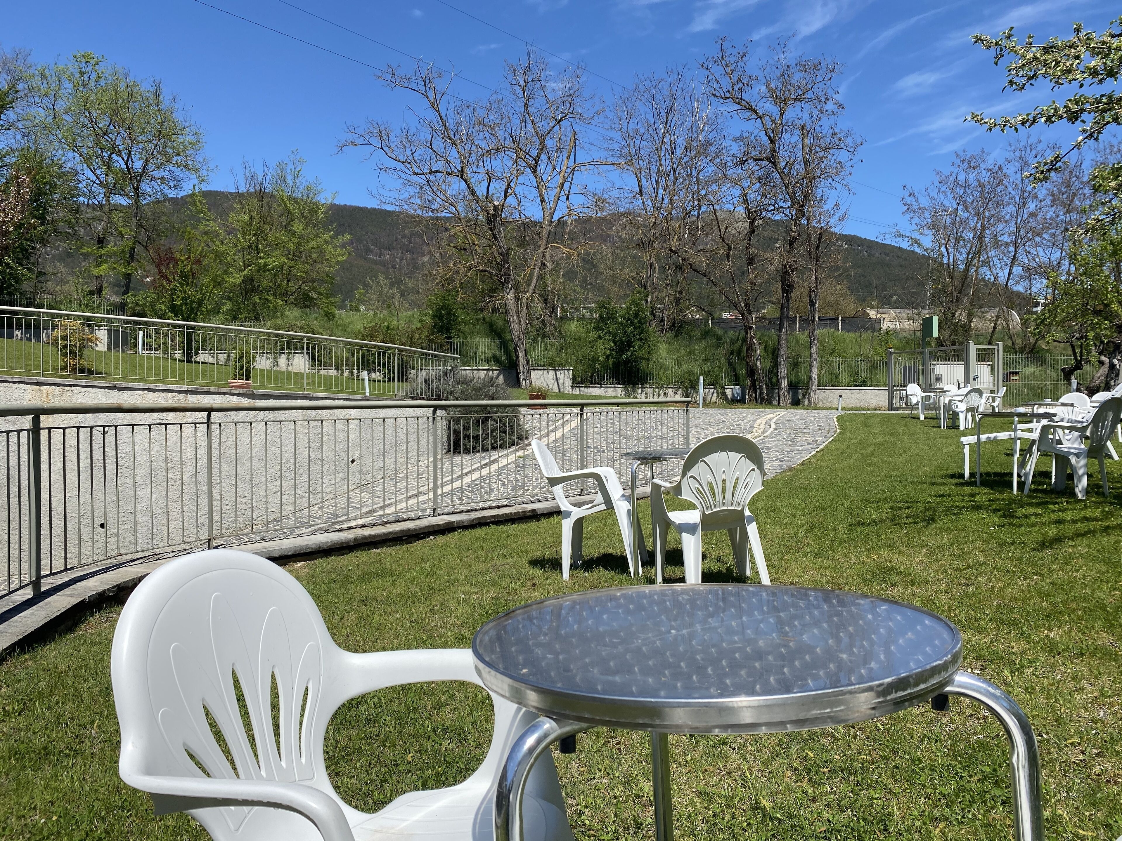 An outdoor seating area with white plastic chairs and tables, surrounded by a metal fence and trees.