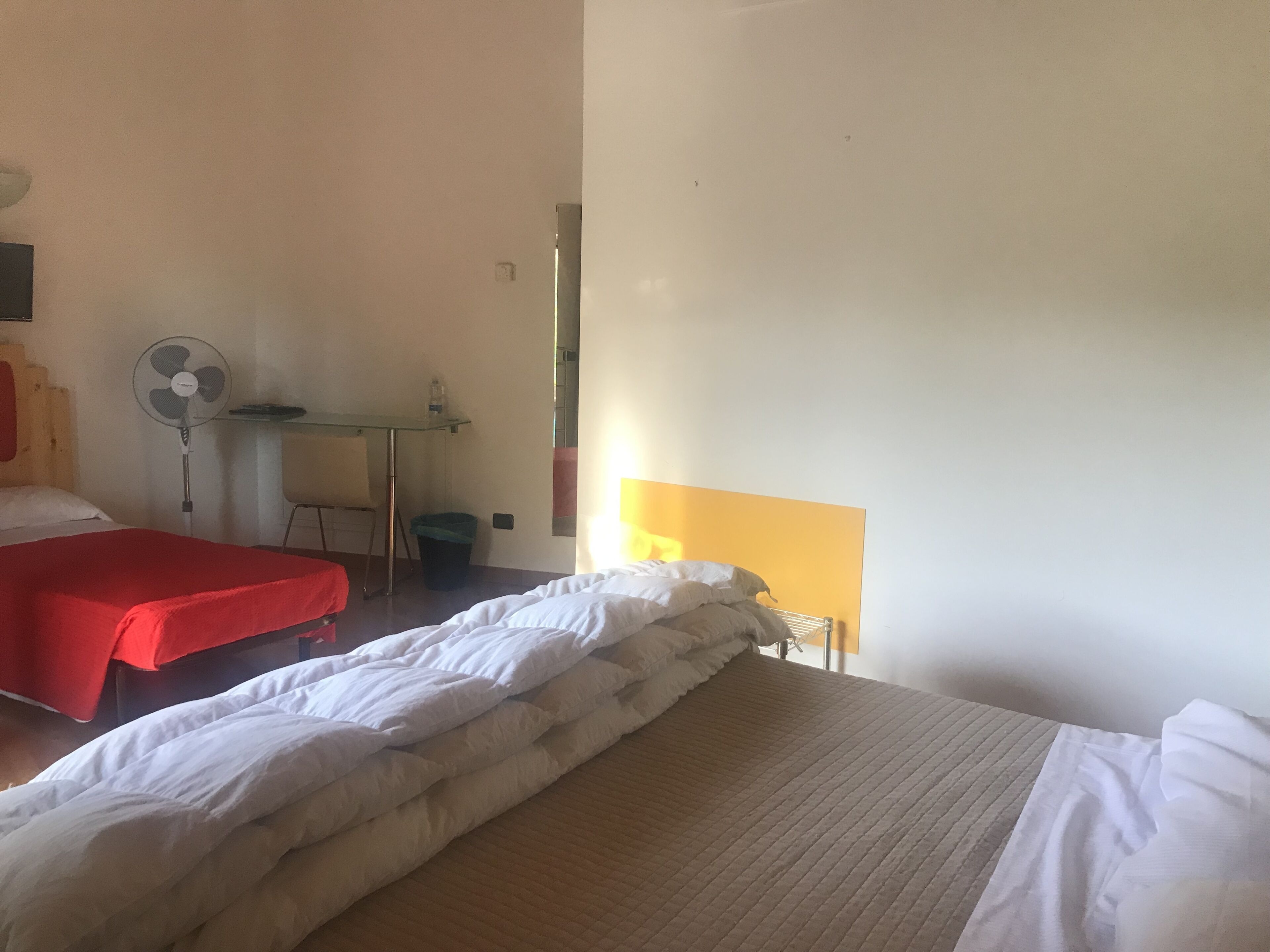 Une chambre d’hôtel avec deux lits, un bureau, une chaise, une poubelle et un ventilateur sur pied.