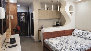 Habitació d'hotel senzilla amb llit, petita cuina, ventilador de sostre i televisió.