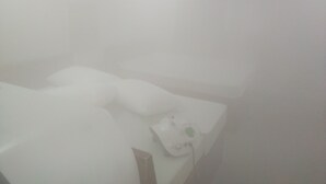 Habitació blanca amb un llit, una tauleta de nit i una taula petita amb una làmpada.