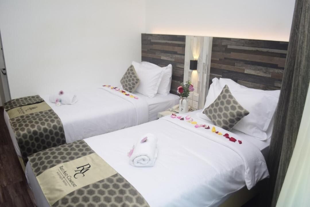 A hotel room with two beds, each with a white bedspread, pillows, and a rolled towel. The room has a wooden headboard and a small table with a vase of flowers.