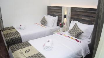 A hotel room with two beds, each with a white bedspread, pillows, and a rolled towel. The room has a wooden headboard and a small table with a vase of flowers.