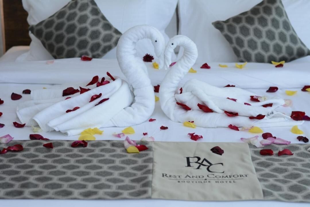 A neatly made bed with a swan-shaped towel arrangement and scattered rose petals. A pillow with a geometric pattern and a tag reading "PAC Rest and Comfort Boutique Hotel" are visible.