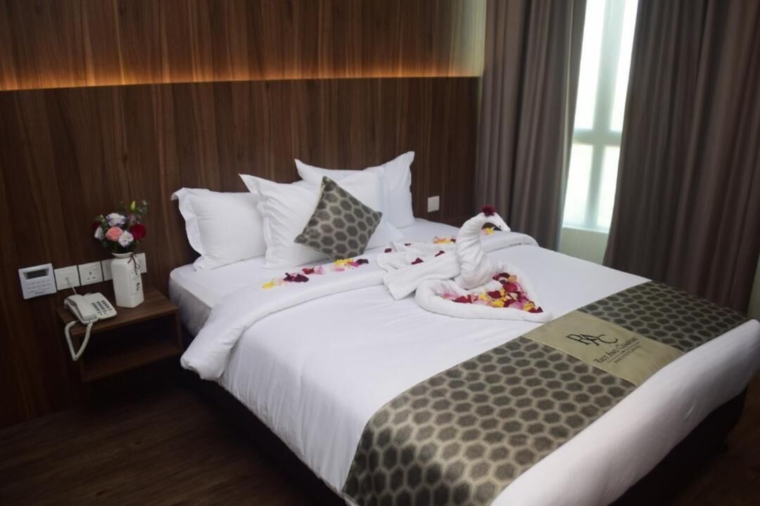 A hotel room with a wooden headboard, a bed with white bedding and a patterned blanket, a bedside table with a phone, and a window with curtains.