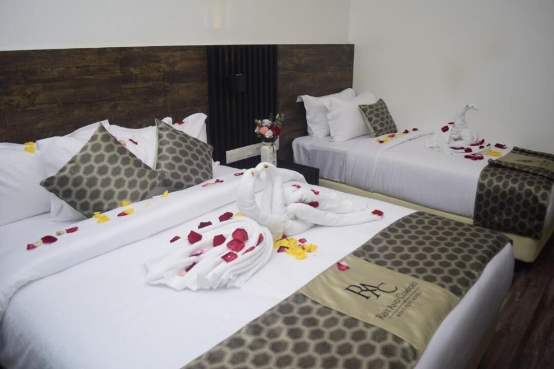 A hotel room with two beds, each with a white bedspread, patterned pillows, and a towel with rose petals. A vase with flowers is on the nightstand.