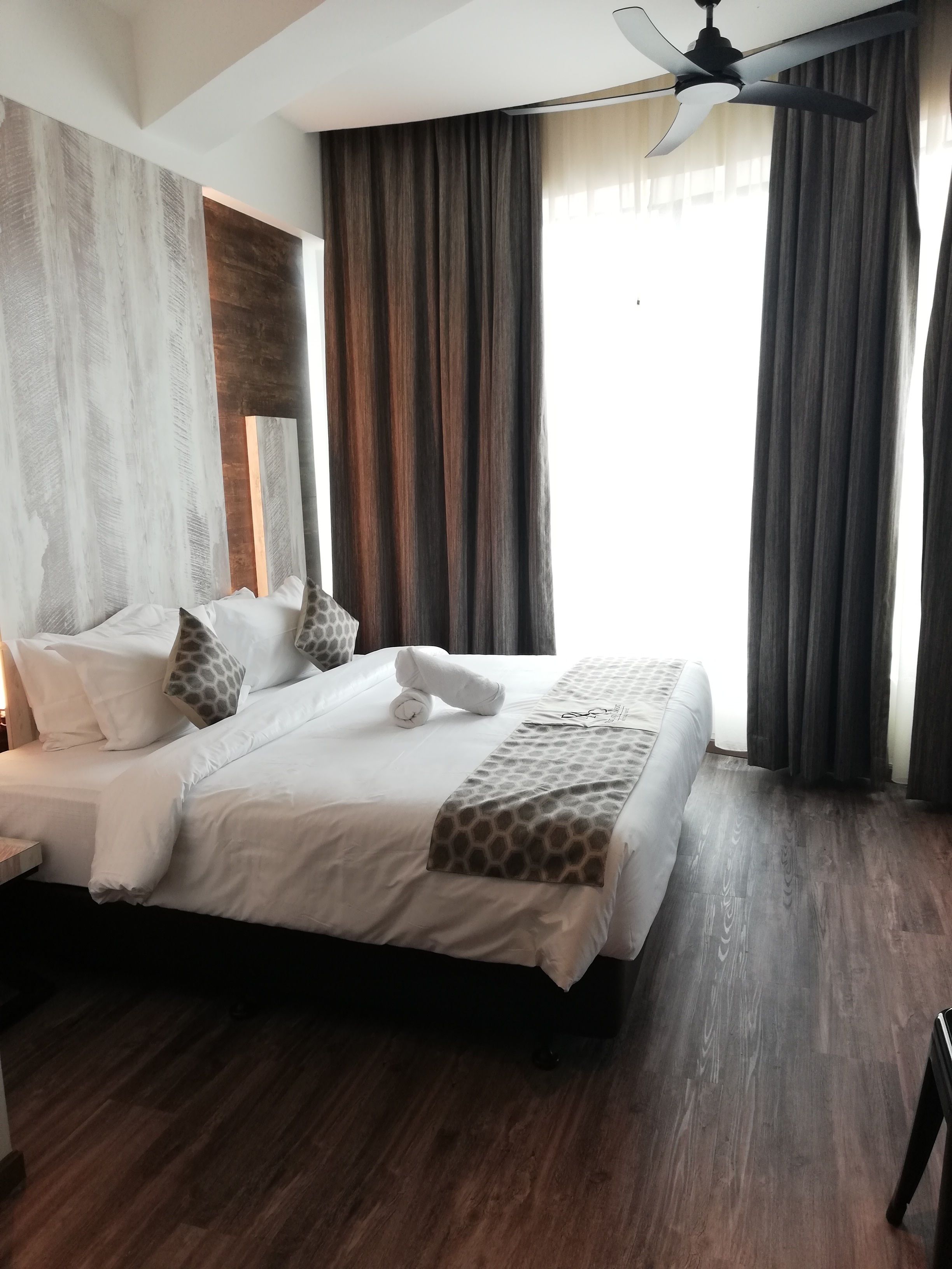 Una cama bien hecha con sábanas blancas y una colcha estampada, un ventilador de techo y cortinas oscuras.