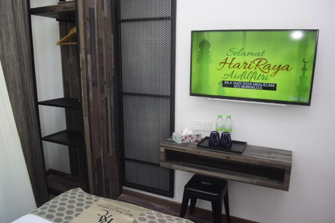 A hotel room with a bed, a wooden wardrobe, a TV mounted on the wall, a small table with a tray, and a shelf with bottles.