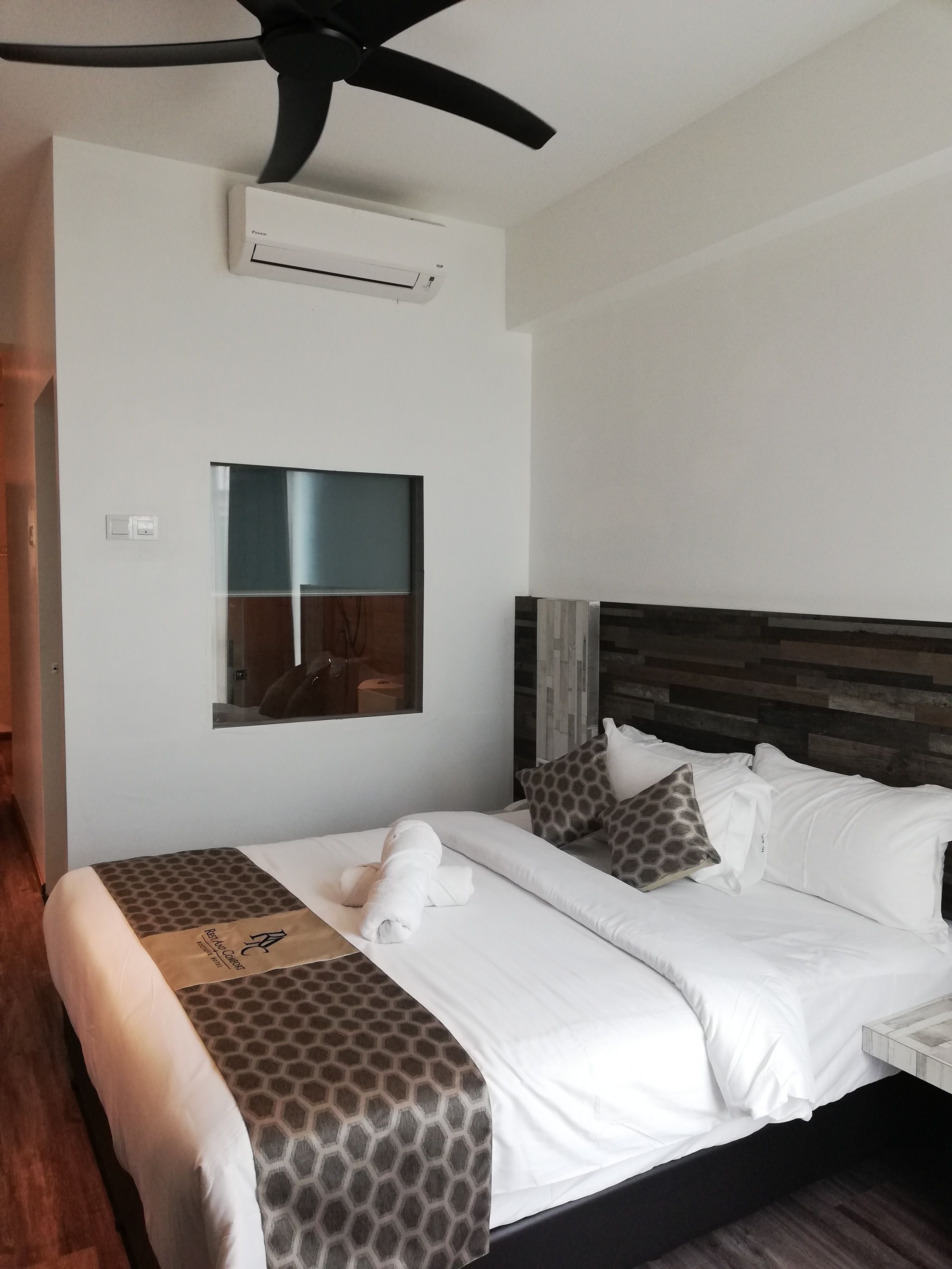 Una camera d'hotel con un letto, una ventola a soffitto, un condizionatore d'aria, una finestra e una testiera in legno.