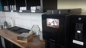 En kaffemaskine med digitalt display, en kaffestation med kopper og et skilt, samt et flisebeklædt bagpanel.