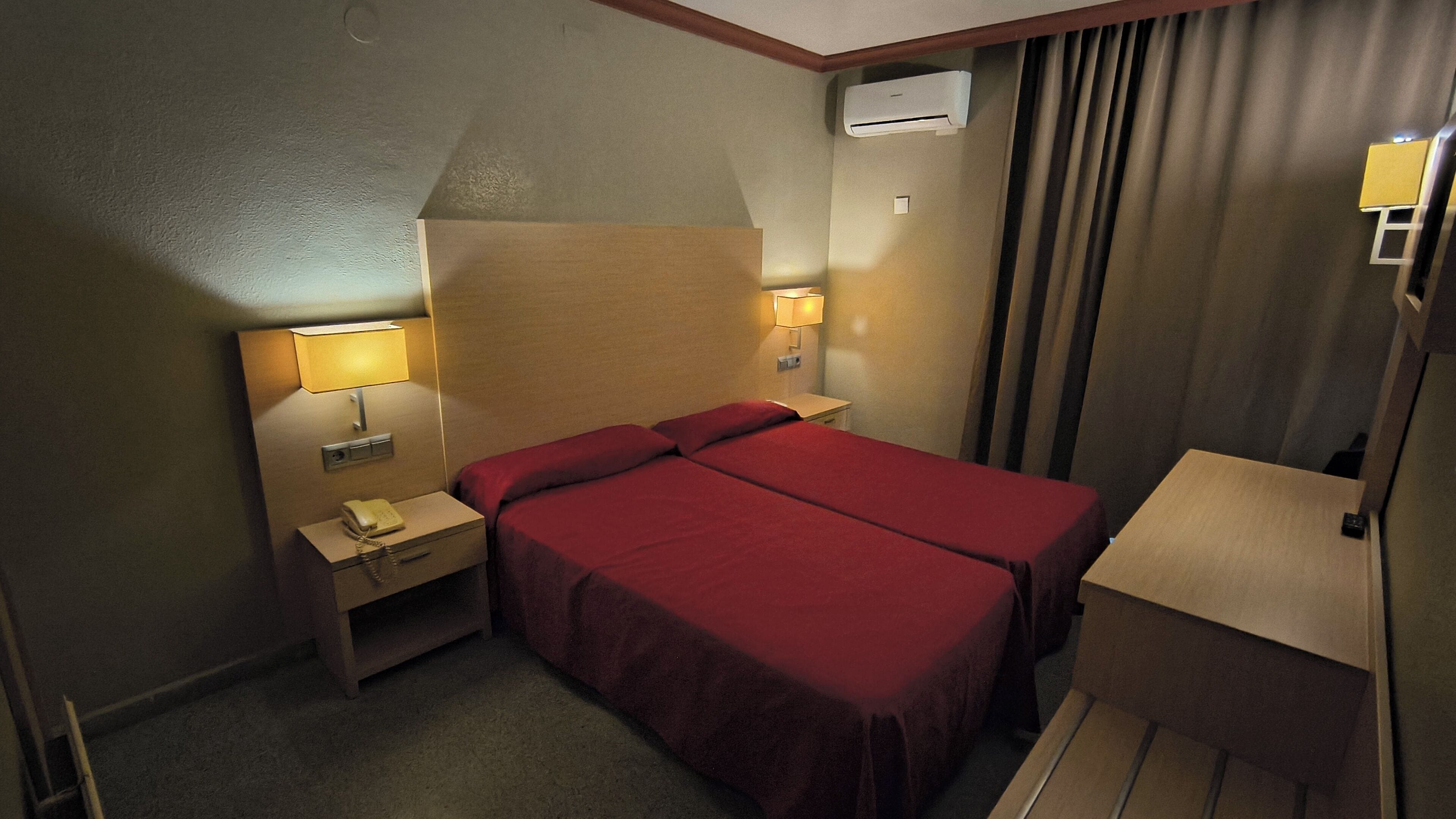 A hotel room with two beds, bedside lamps, a telephone, an air conditioning unit, and a desk with a lamp.