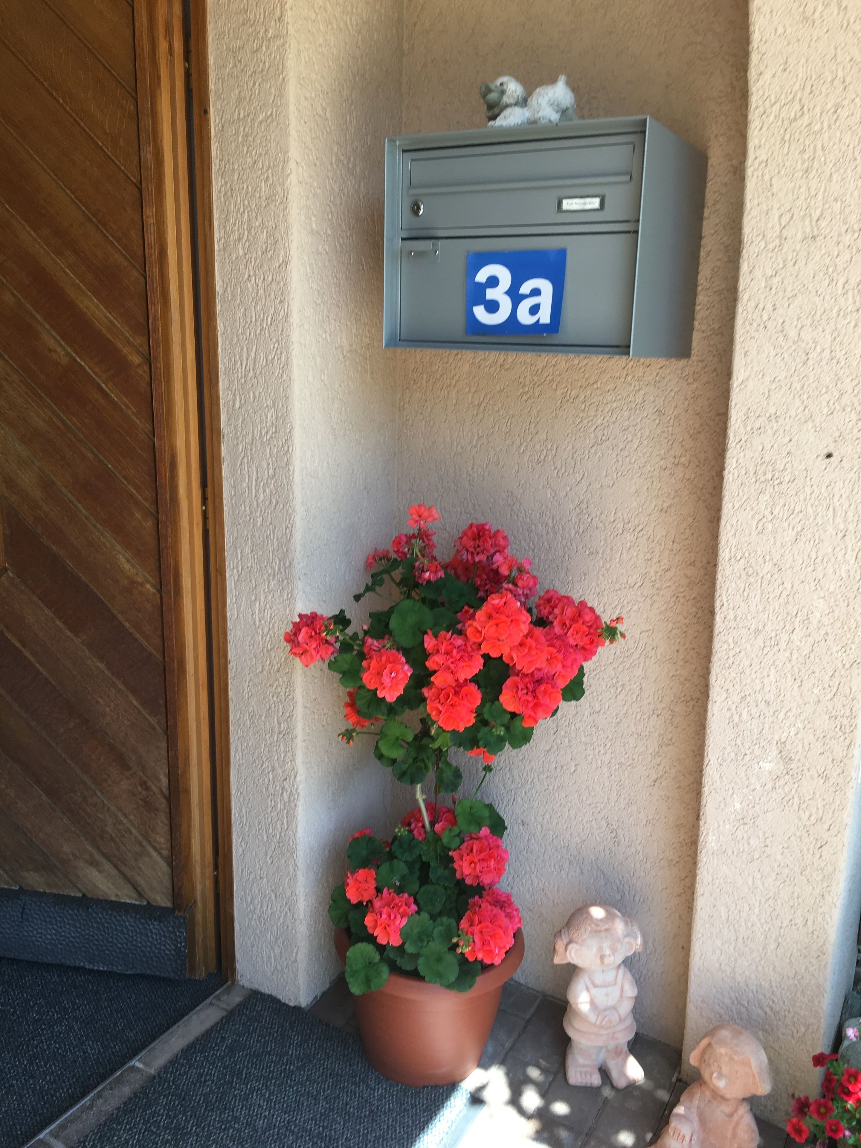 A mailbox labeled '3a' mounted on a wall, a potted red flower plant, and a small statue of a child.